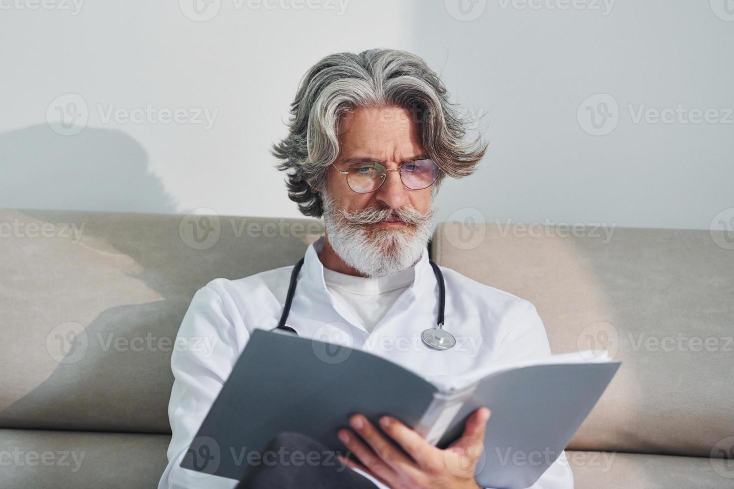 Senior male doctor with grey hair and beard in white coat is indoors in clinic sits on sofa with notepad in hands photo