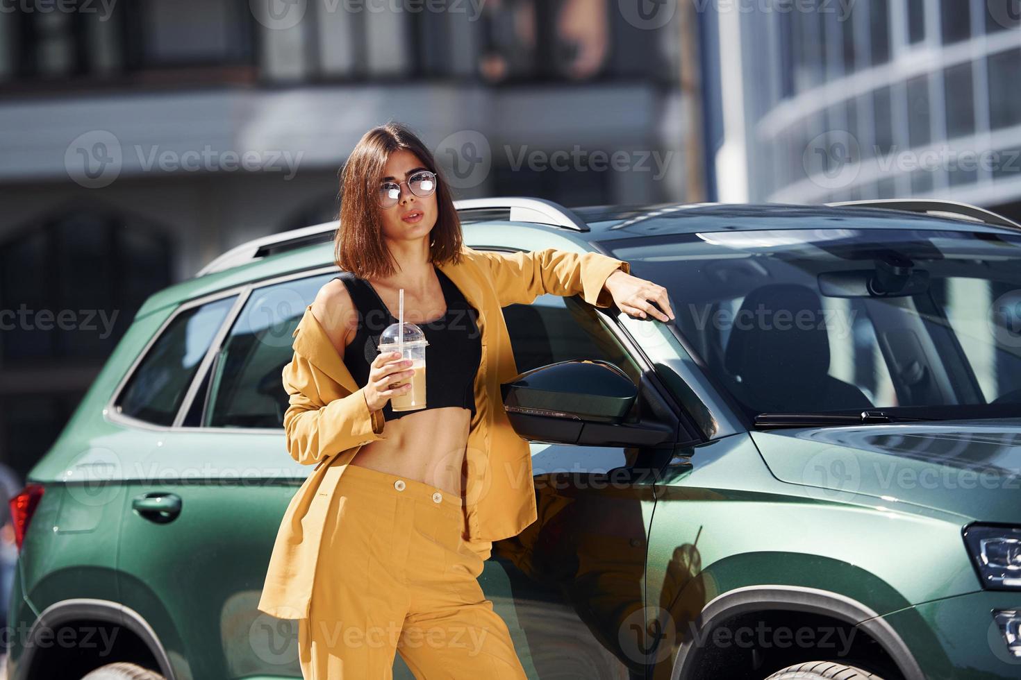 With drink in hands. Young fashionable woman in burgundy colored coat at daytime with her car photo