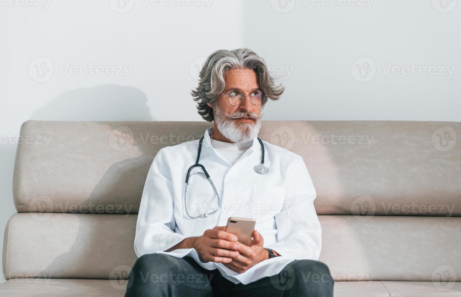 Senior male doctor with grey hair and beard in white coat is indoors in clinic sits on sofa with phone in hands photo