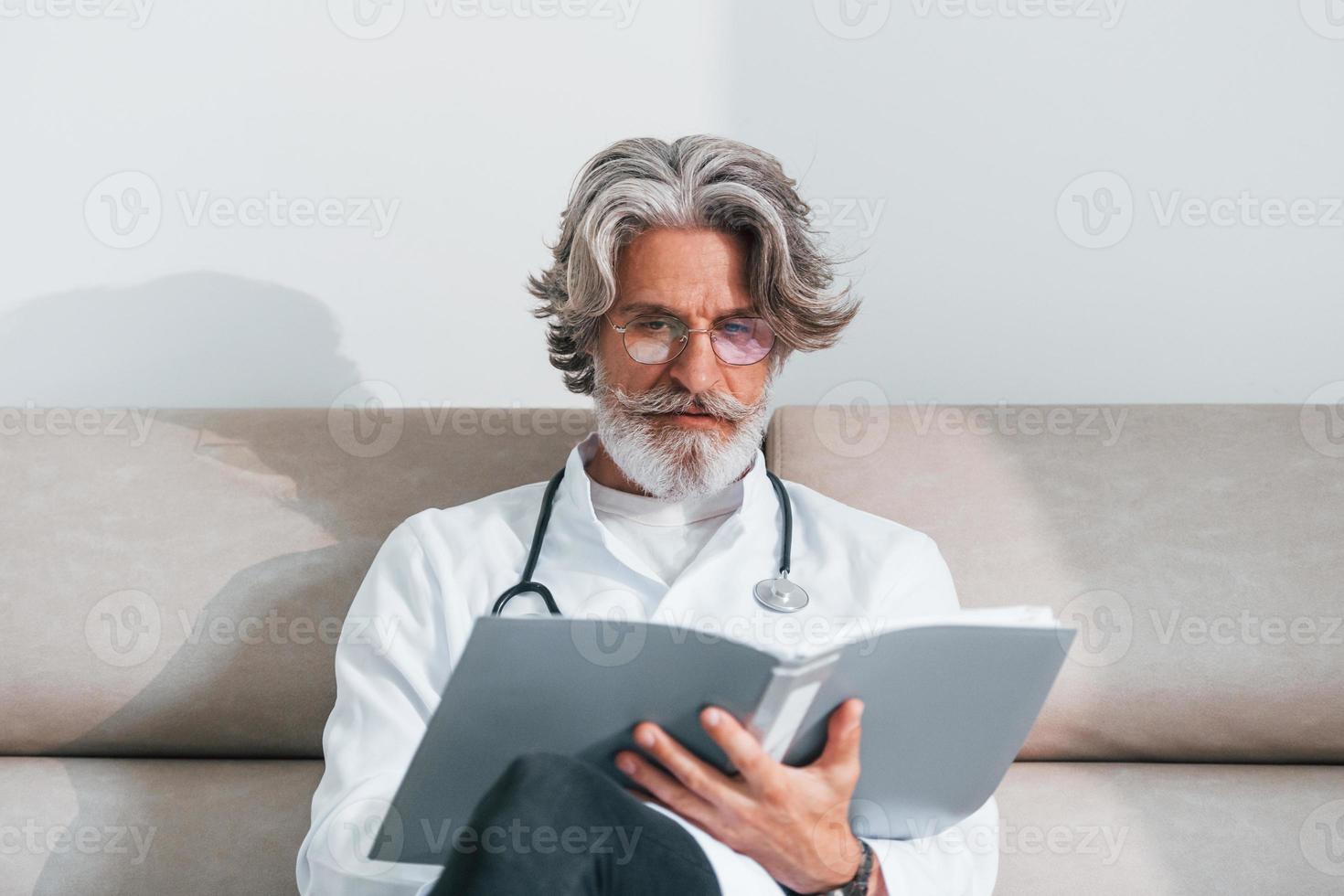 Senior male doctor with grey hair and beard in white coat is indoors in clinic sits on sofa with notepad in hands photo