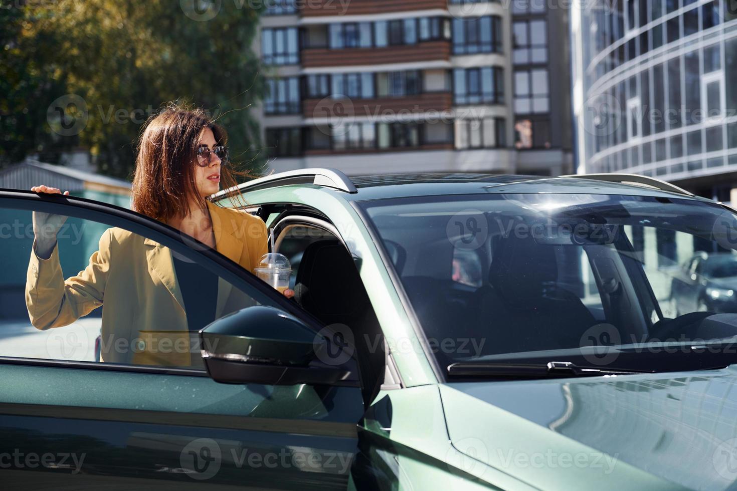 Against business building. Young fashionable woman in burgundy colored coat at daytime with her car photo