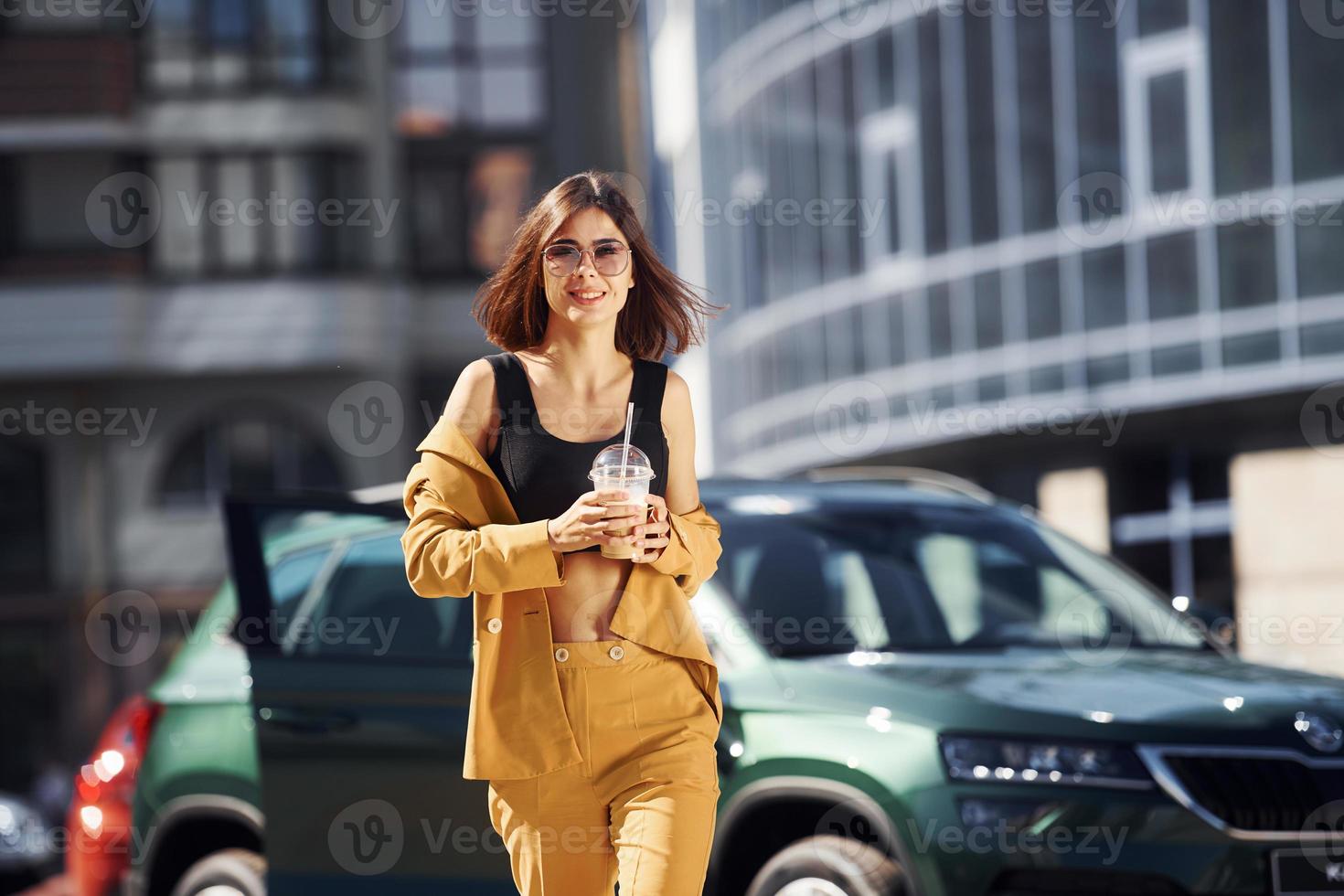 Against business building. Young fashionable woman in burgundy colored coat at daytime with her car photo