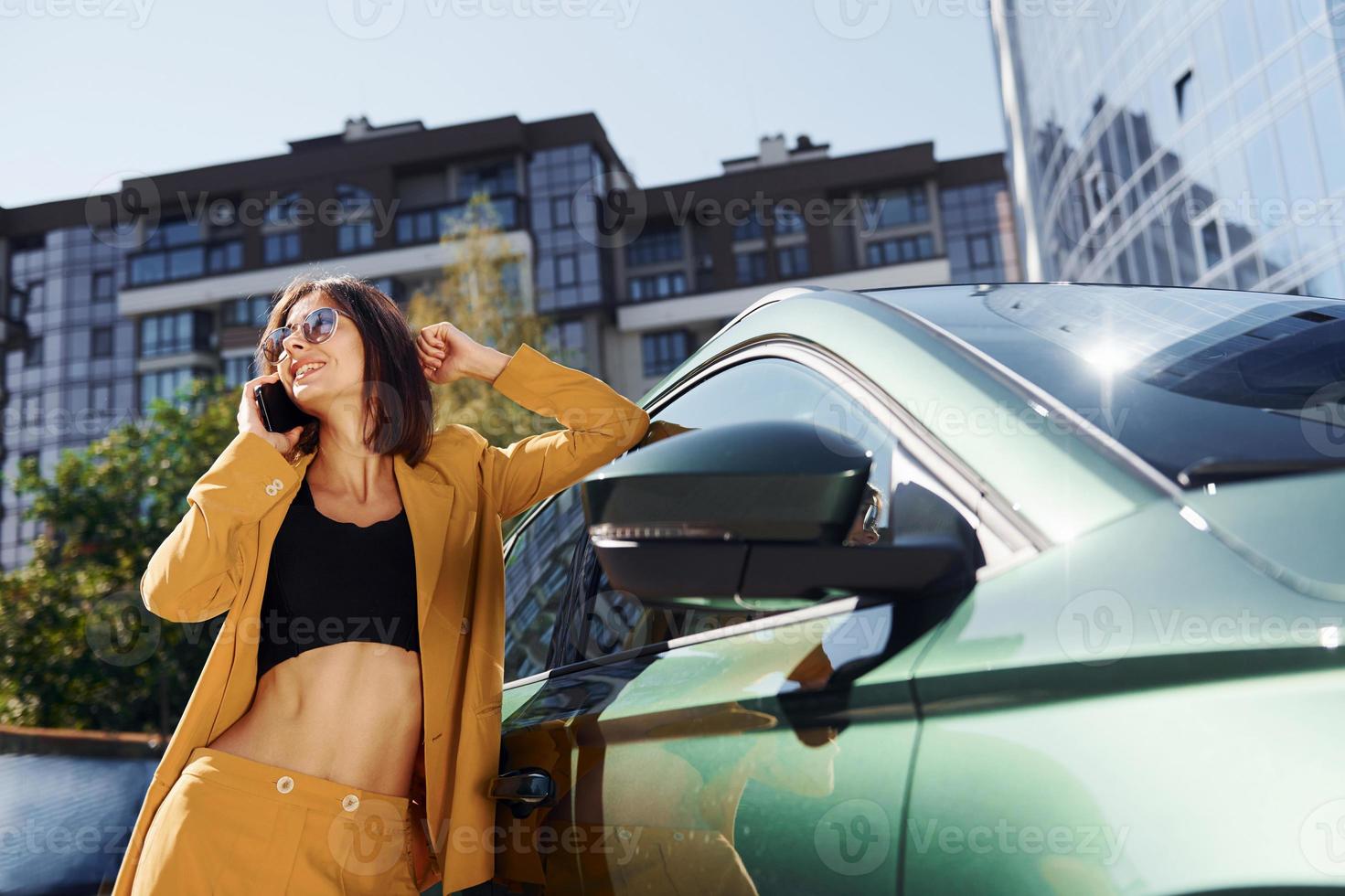 With smartphone. Young fashionable woman in burgundy colored coat at daytime with her car photo