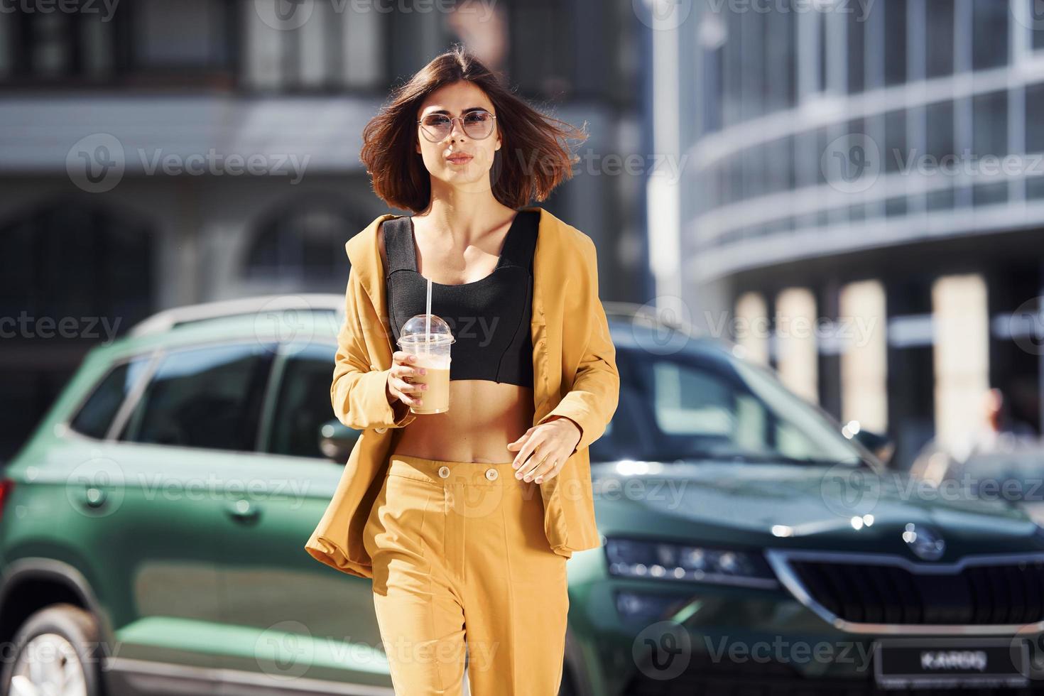 With drink in hands. Young fashionable woman in burgundy colored coat at daytime with her car photo