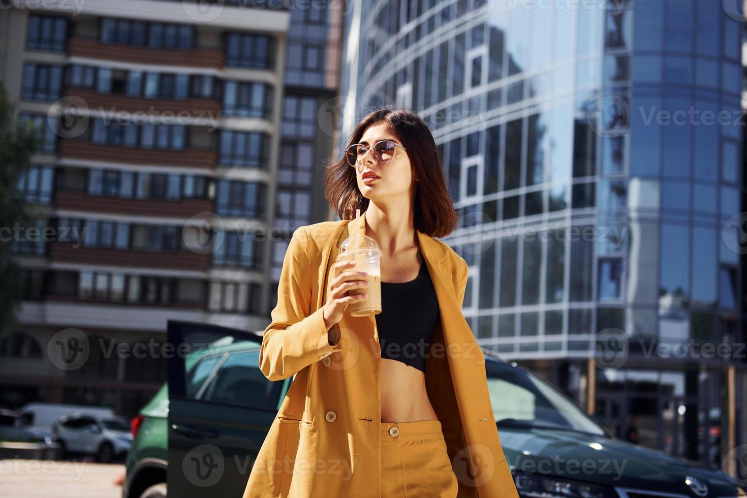 Against business building. Young fashionable woman in burgundy colored coat at daytime with her car photo