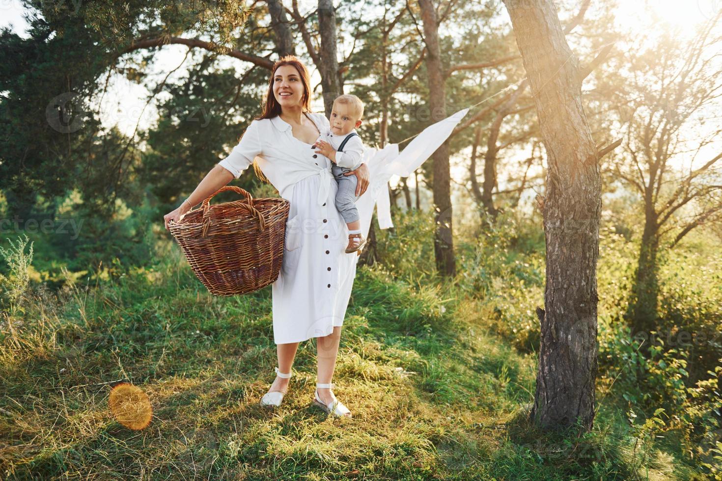 Positive housewife with hangs white clothes to dry. Young mother with her little son is outdoors in the forest. Beautiful sunshine photo