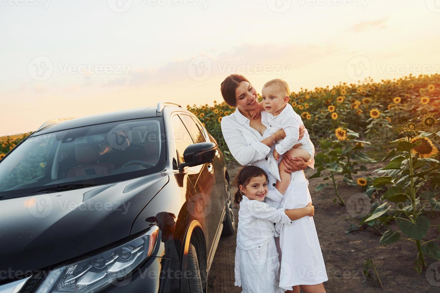 Near modern black car. Young mother with her little son and daughter is outdoors in the agricultural field. Beautiful sunshine photo