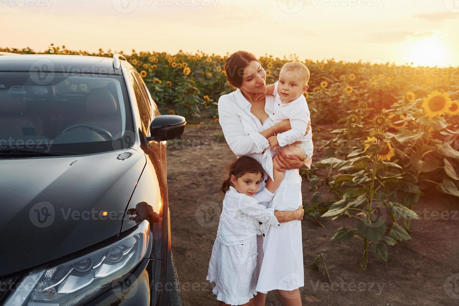 cerca de un coche negro moderno. la joven madre con su pequeño hijo y su hija está al aire libre en el campo agrícola. hermoso amanecer foto