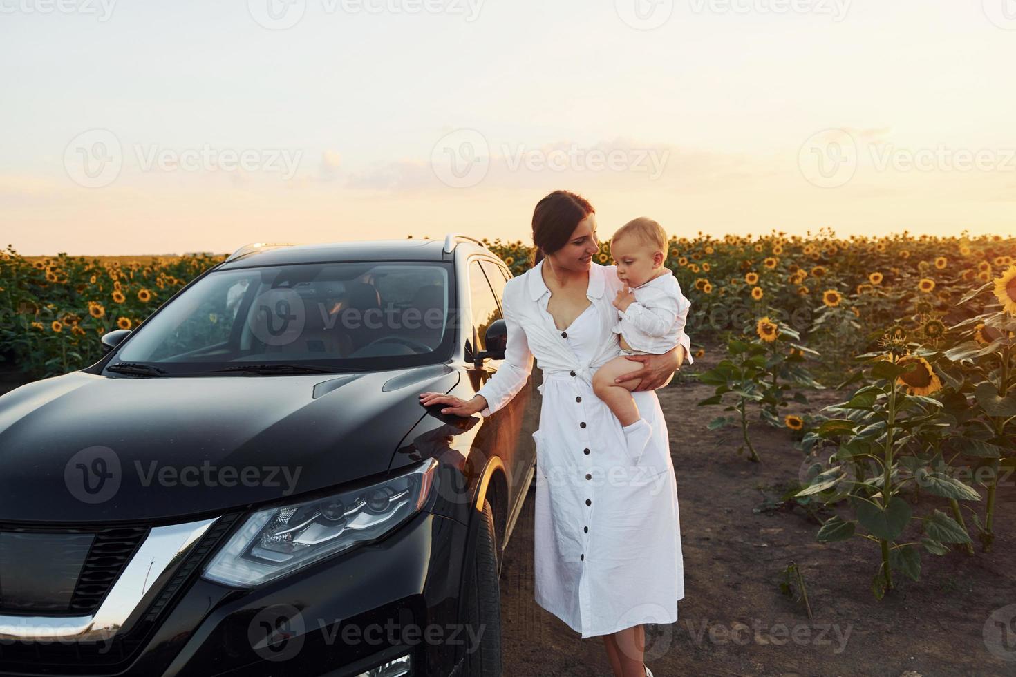 cerca de un coche negro moderno. la joven madre con su hijo pequeño está al aire libre en el campo agrícola. hermoso amanecer foto