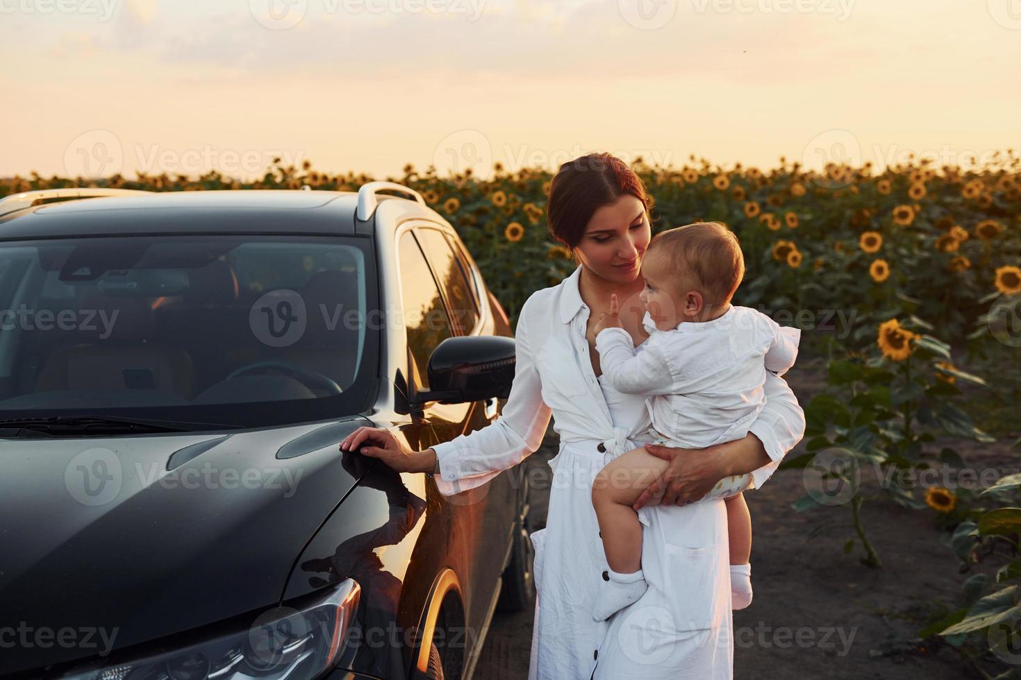 cerca de un coche negro moderno. la joven madre con su hijo pequeño está al aire libre en el campo agrícola. hermoso amanecer foto