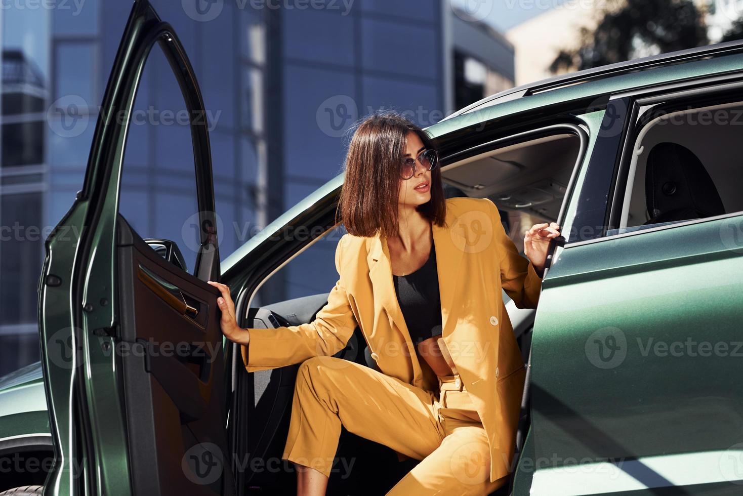 Exits vehicle. Young fashionable woman in burgundy colored coat at daytime with her car photo