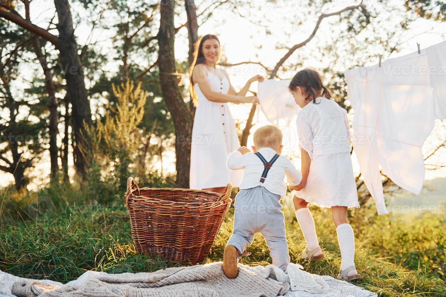 White clothes hanging on the rope to dry. Young mother with her little daughter and son is outdoors in the forest. Beautiful sunshine photo