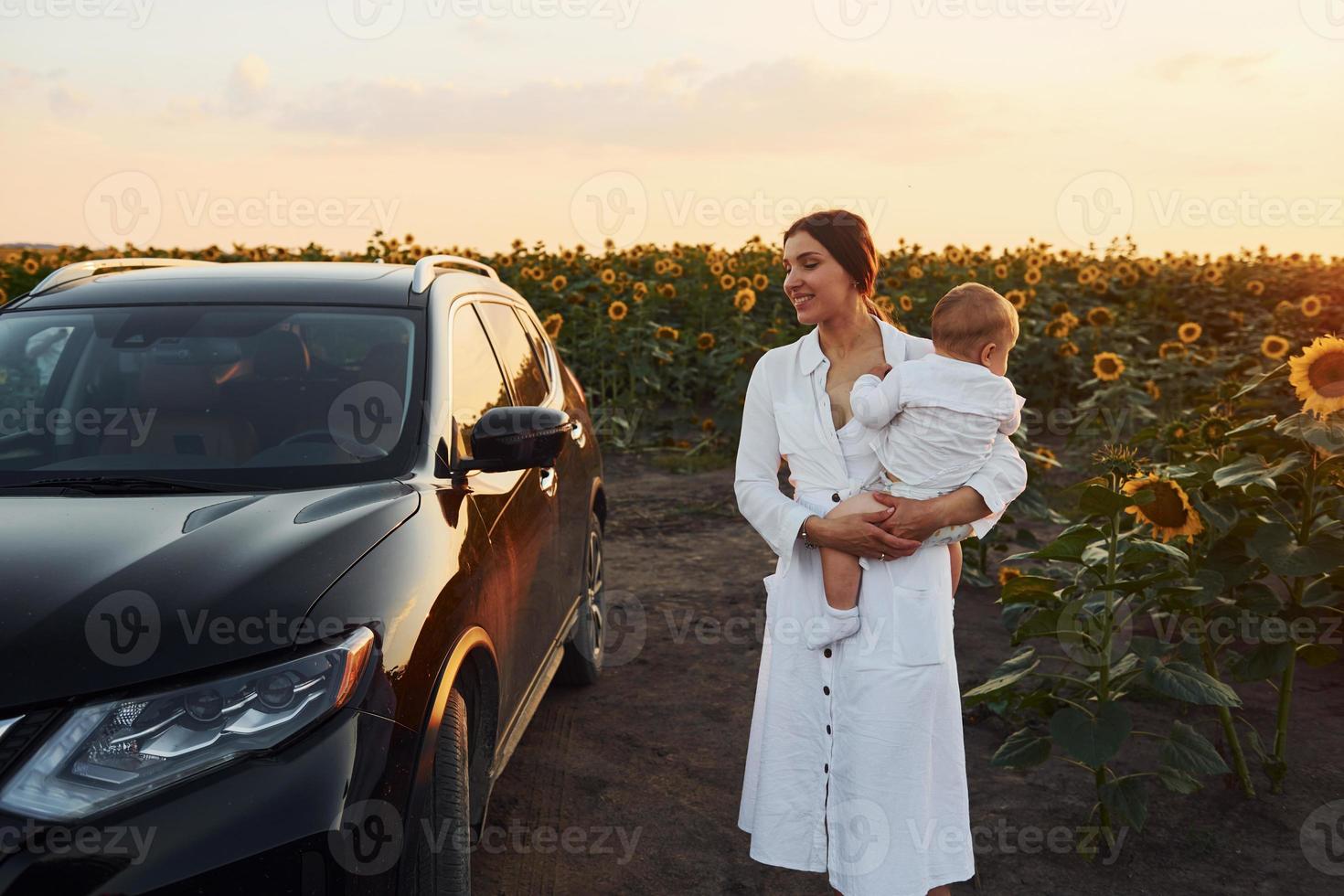 cerca de un coche negro moderno. la joven madre con su hijo pequeño está al aire libre en el campo agrícola. hermoso amanecer foto