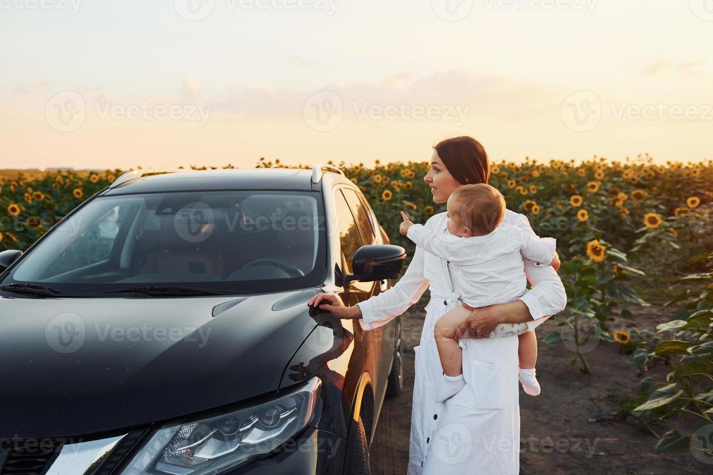 cerca de un coche negro moderno. la joven madre con su hijo pequeño está al aire libre en el campo agrícola. hermoso amanecer foto