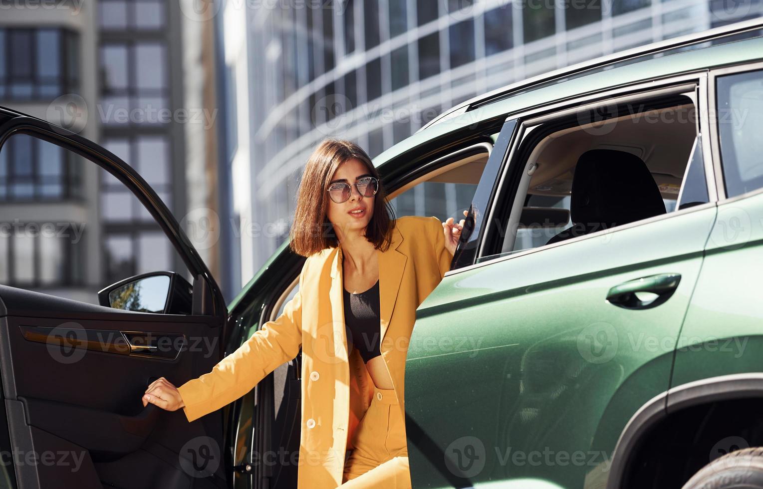 Exits vehicle. Young fashionable woman in burgundy colored coat at daytime with her car photo