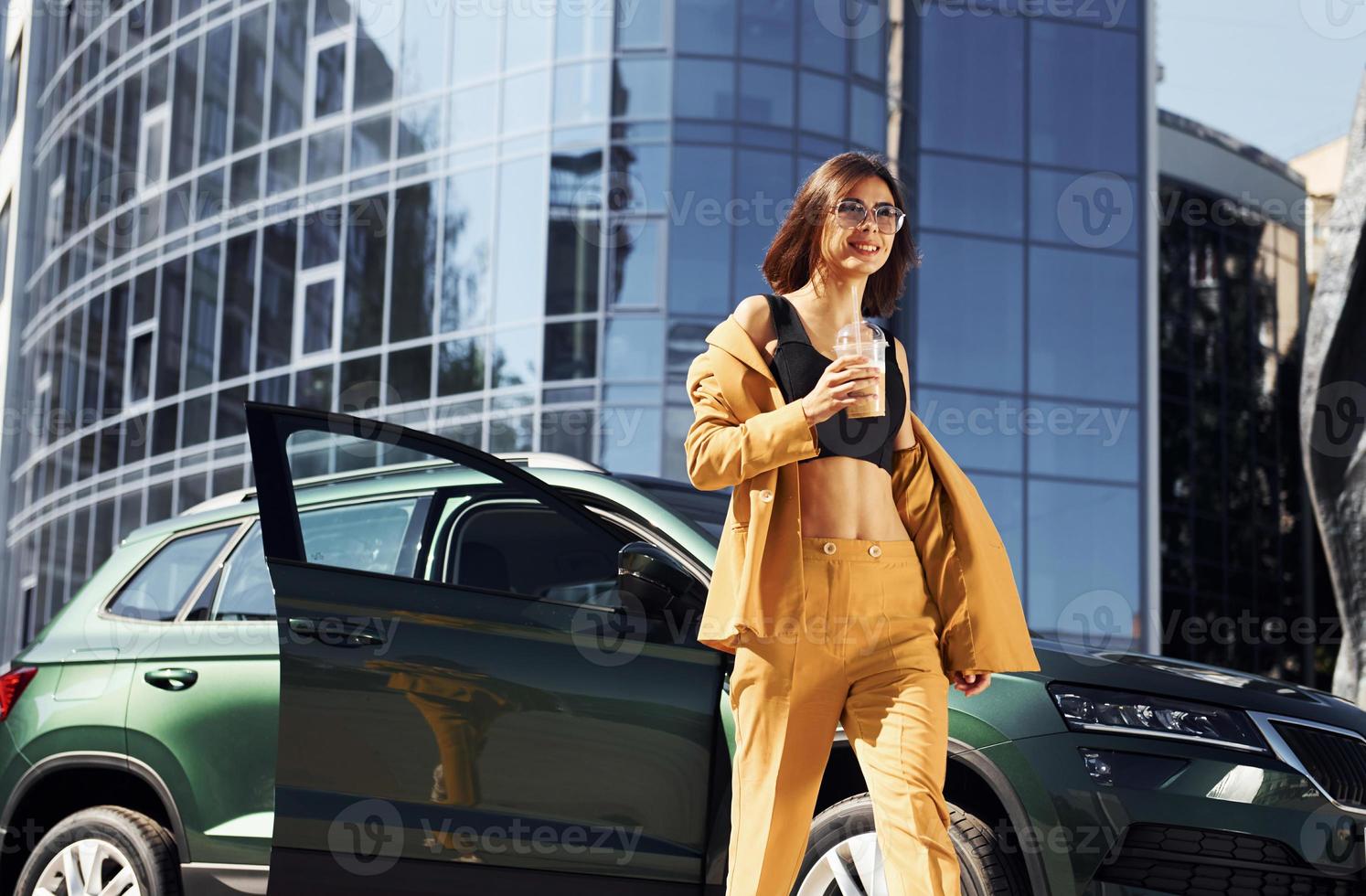 Takes break with cup of coffee. Young fashionable woman in burgundy colored coat at daytime with her car photo