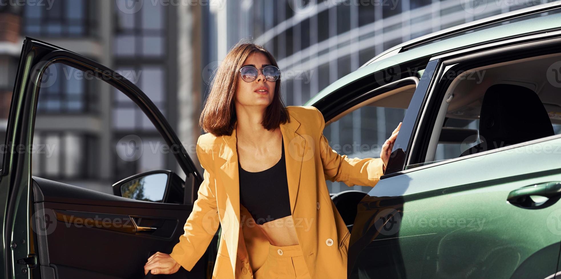 Exits vehicle. Young fashionable woman in burgundy colored coat at daytime with her car photo
