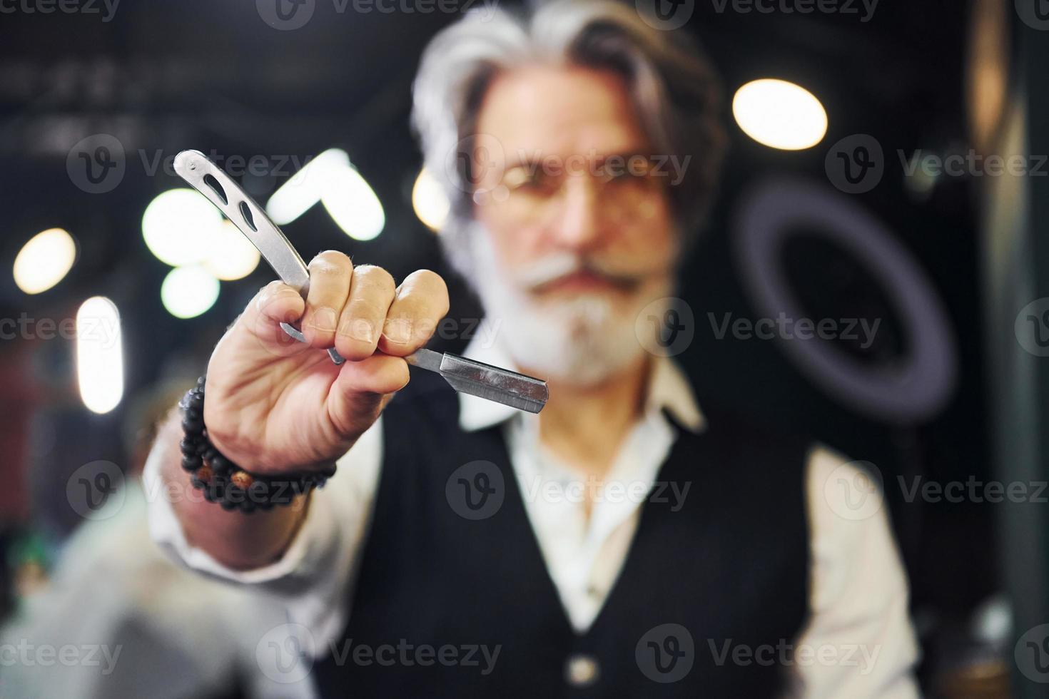 Holds barbershop equipment. Stylish modern senior man with gray hair and beard is indoors photo