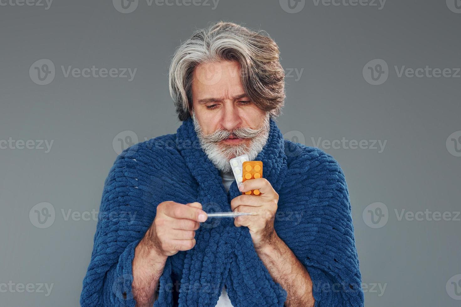 Getting sick. Against grey background. Stylish modern senior man with gray hair and beard is indoors photo