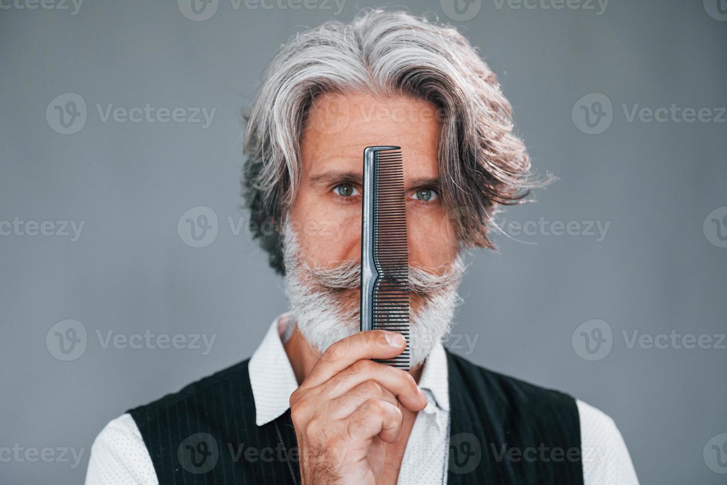 Professional barbershop worker. Against grey background. Stylish modern senior man with gray hair and beard is indoors photo