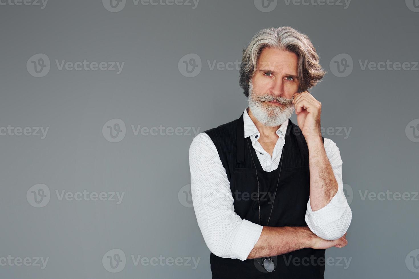 Against grey background. Stylish modern senior man with gray hair and beard is indoors photo