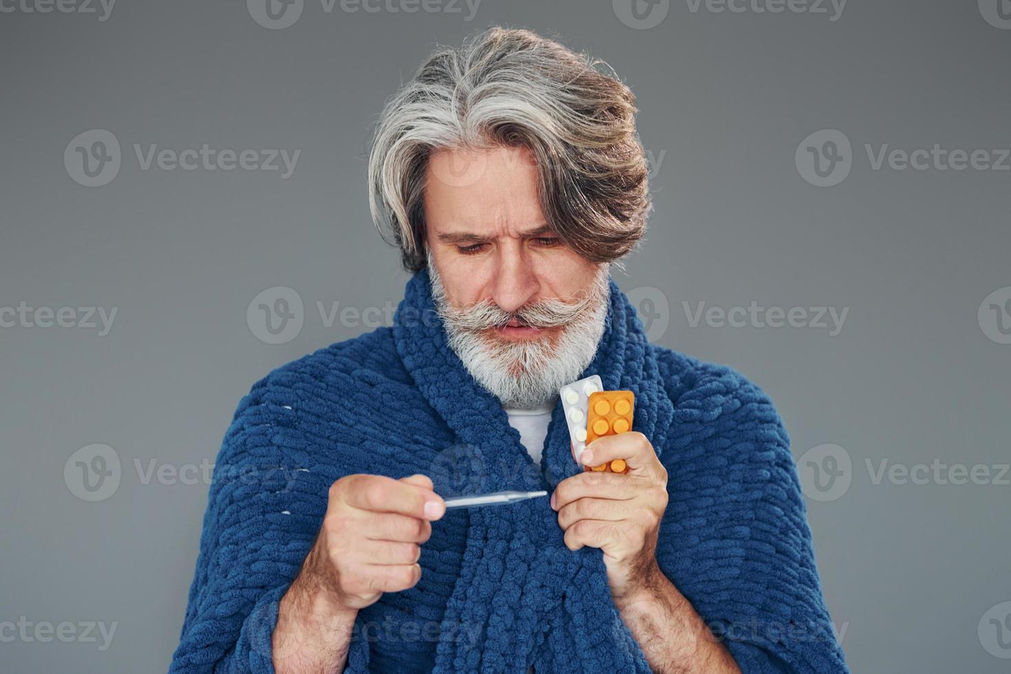 Getting sick. Against grey background. Stylish modern senior man with gray hair and beard is indoors photo