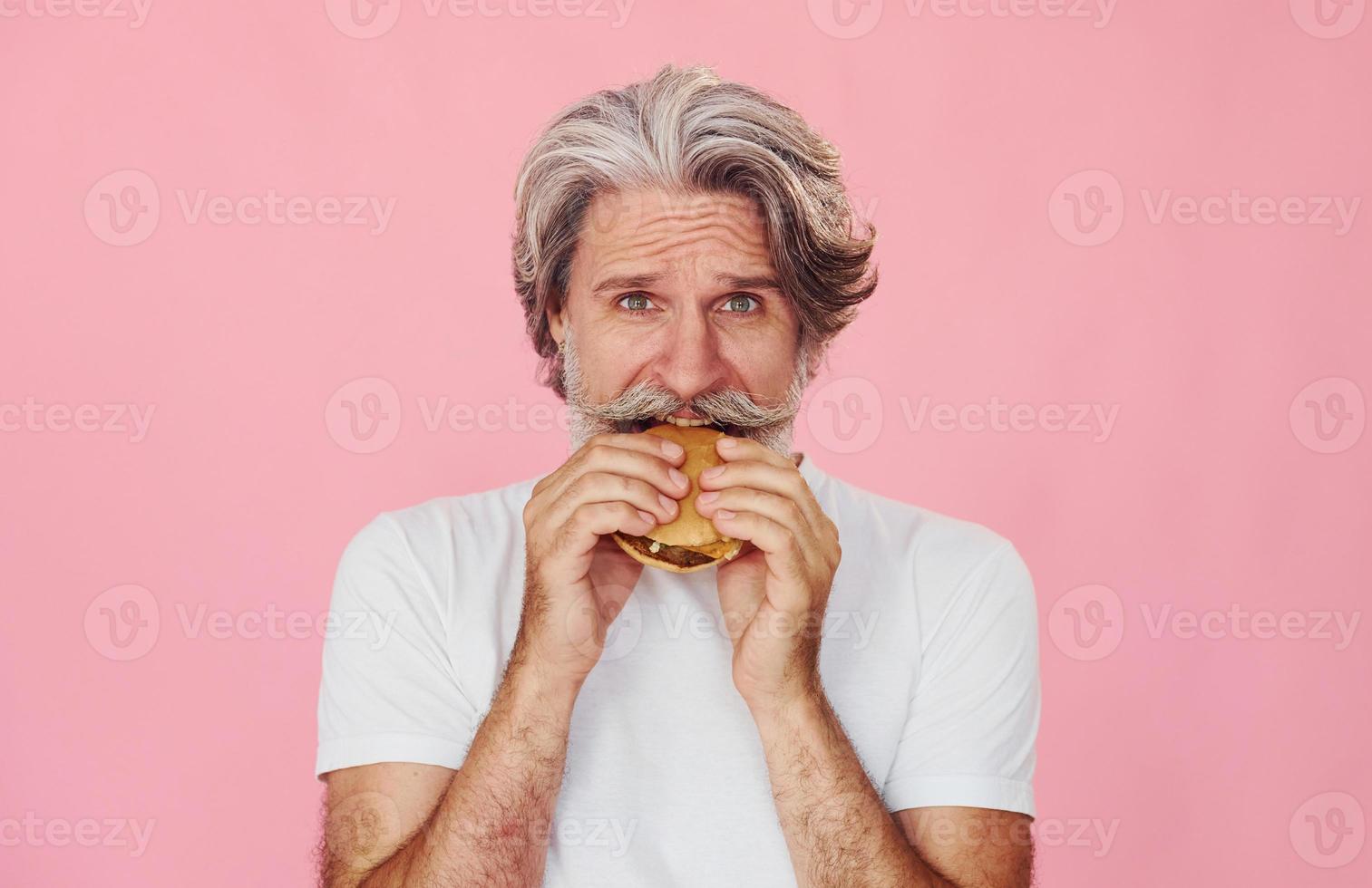 Eats delicious burger. Stylish modern senior man with gray hair and beard is indoors photo