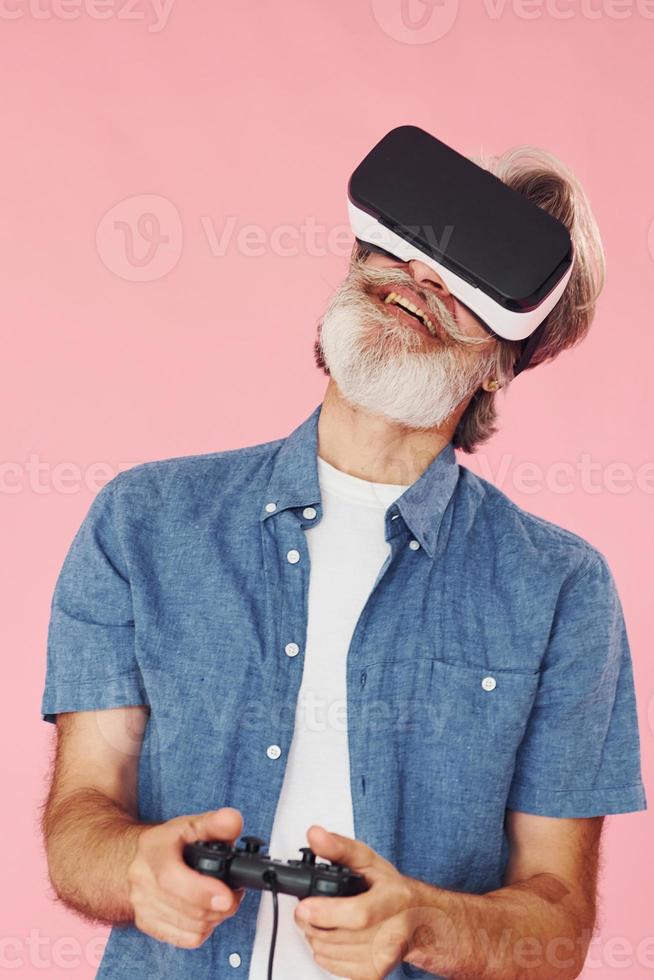 Playing game with virtual reality glasses and joystick. Stylish modern senior man with gray hair and beard is indoors photo