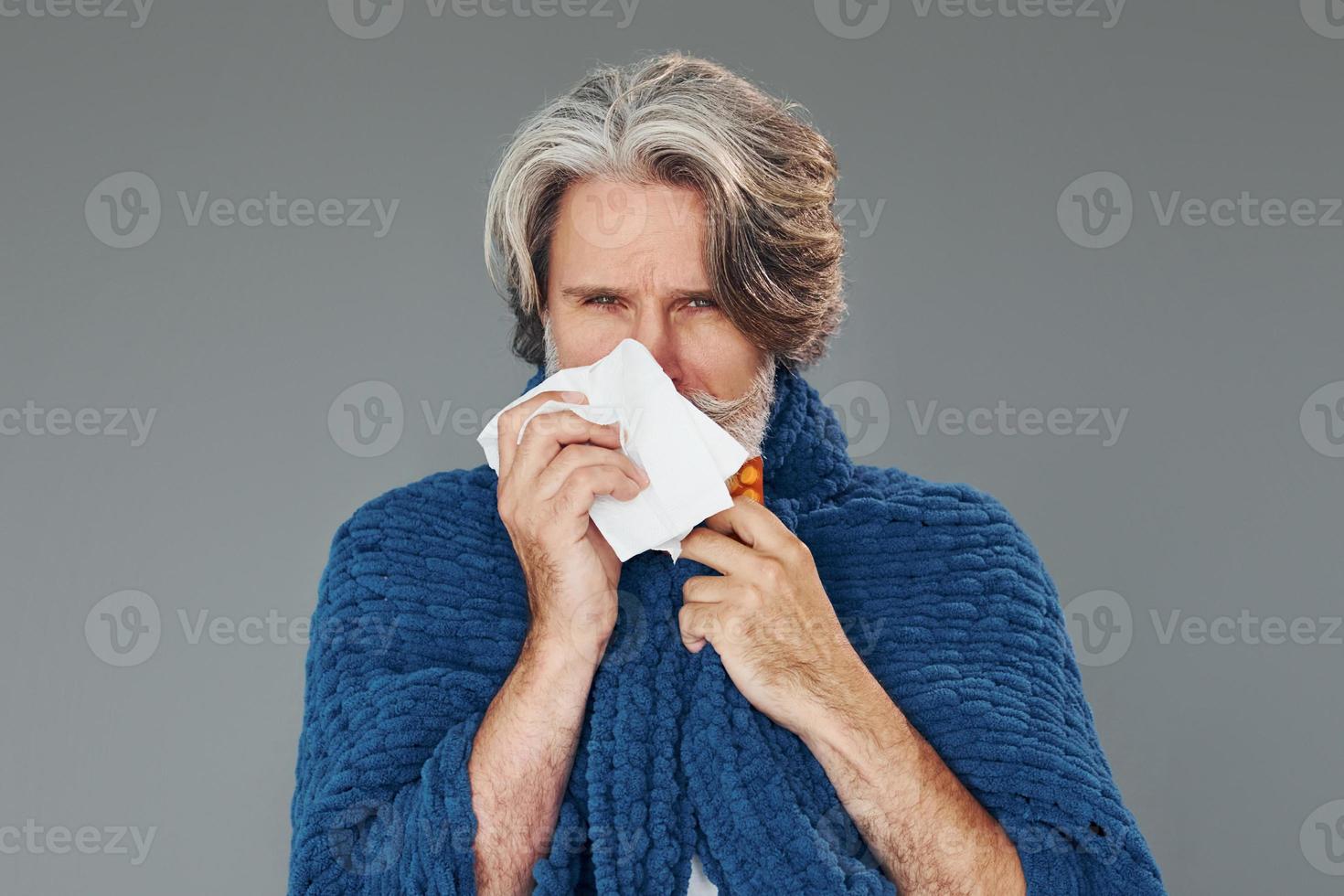 Getting sick. Against grey background. Stylish modern senior man with gray hair and beard is indoors photo