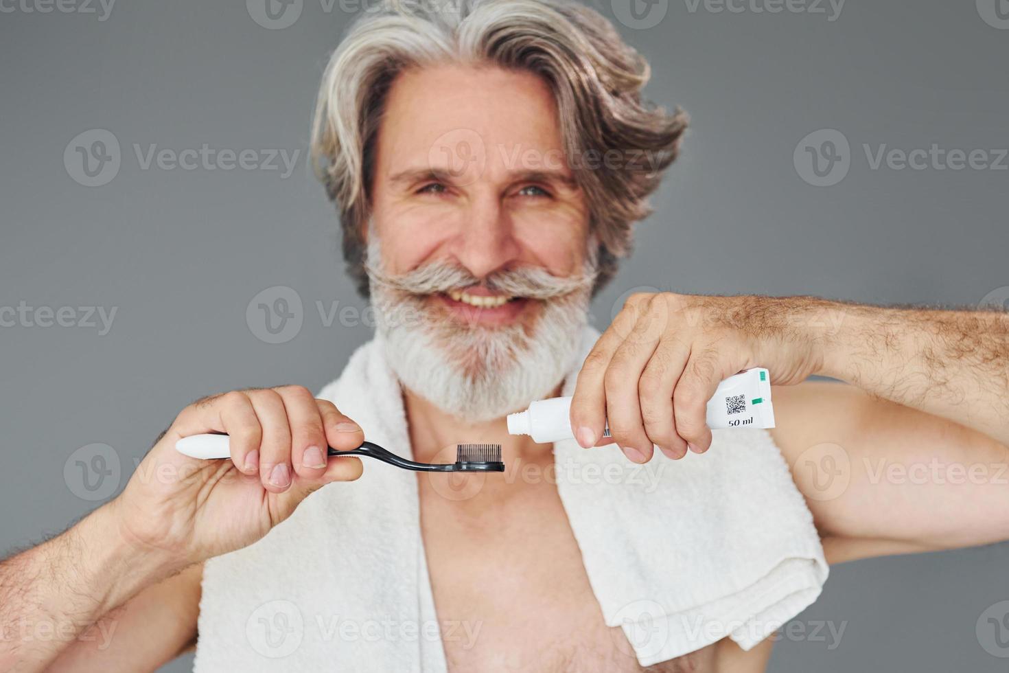 Using toothpaste. Stylish modern senior man with gray hair and beard is