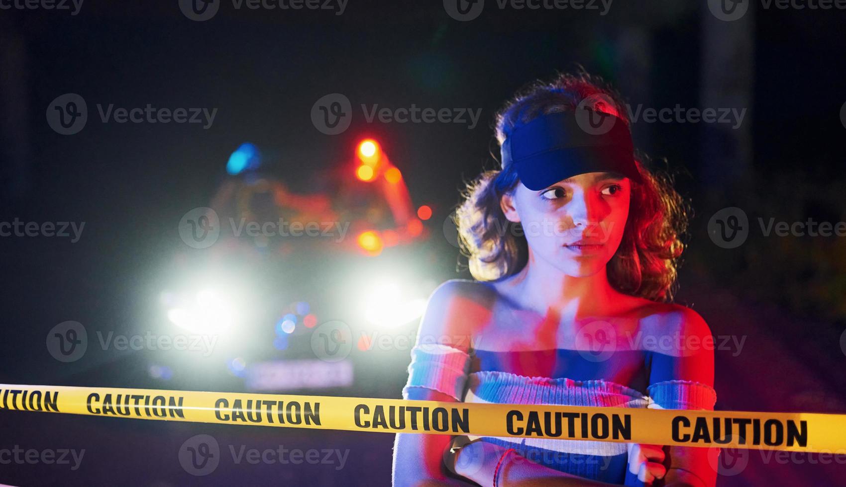 Cute young girl with curly hair and in hat standing outdoors near police line tape. Conception of crime or riot photo