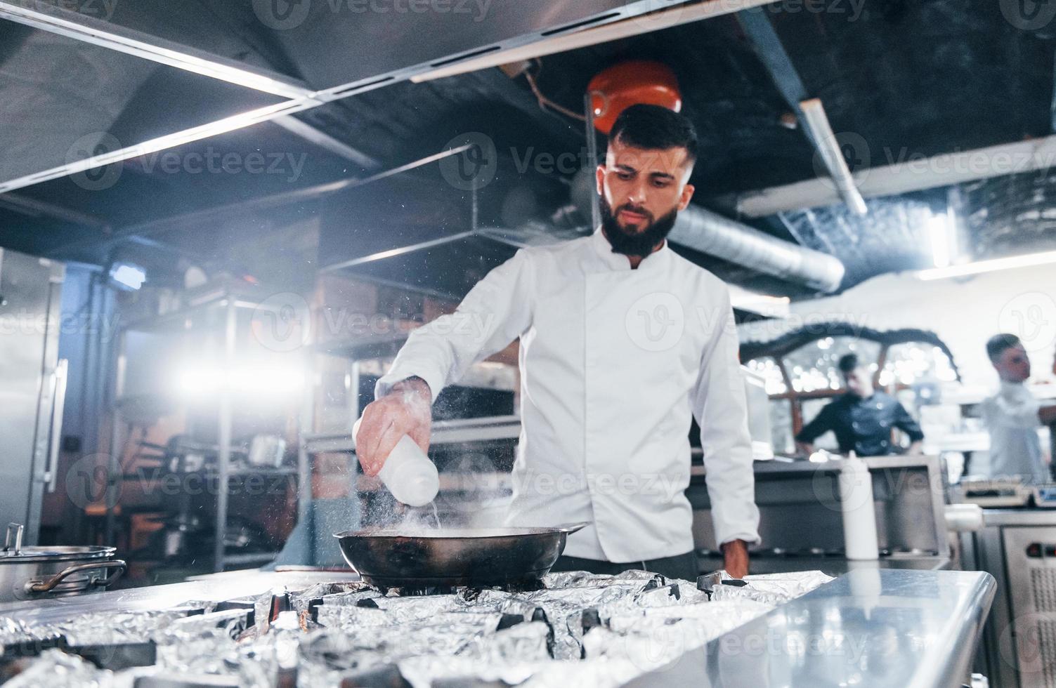 Uses frying pan. Chef in white uniform cooking food at kitchen. Busy day at work photo