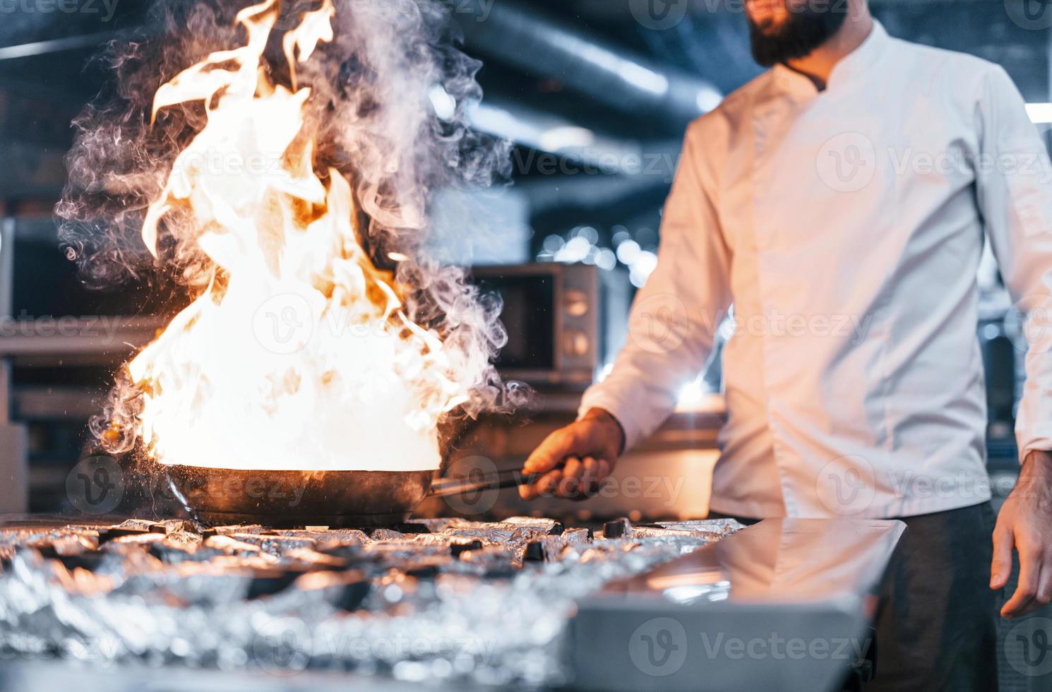 Frying pan is on fire. Chef in white uniform cooking food at kitchen. Busy day at work photo