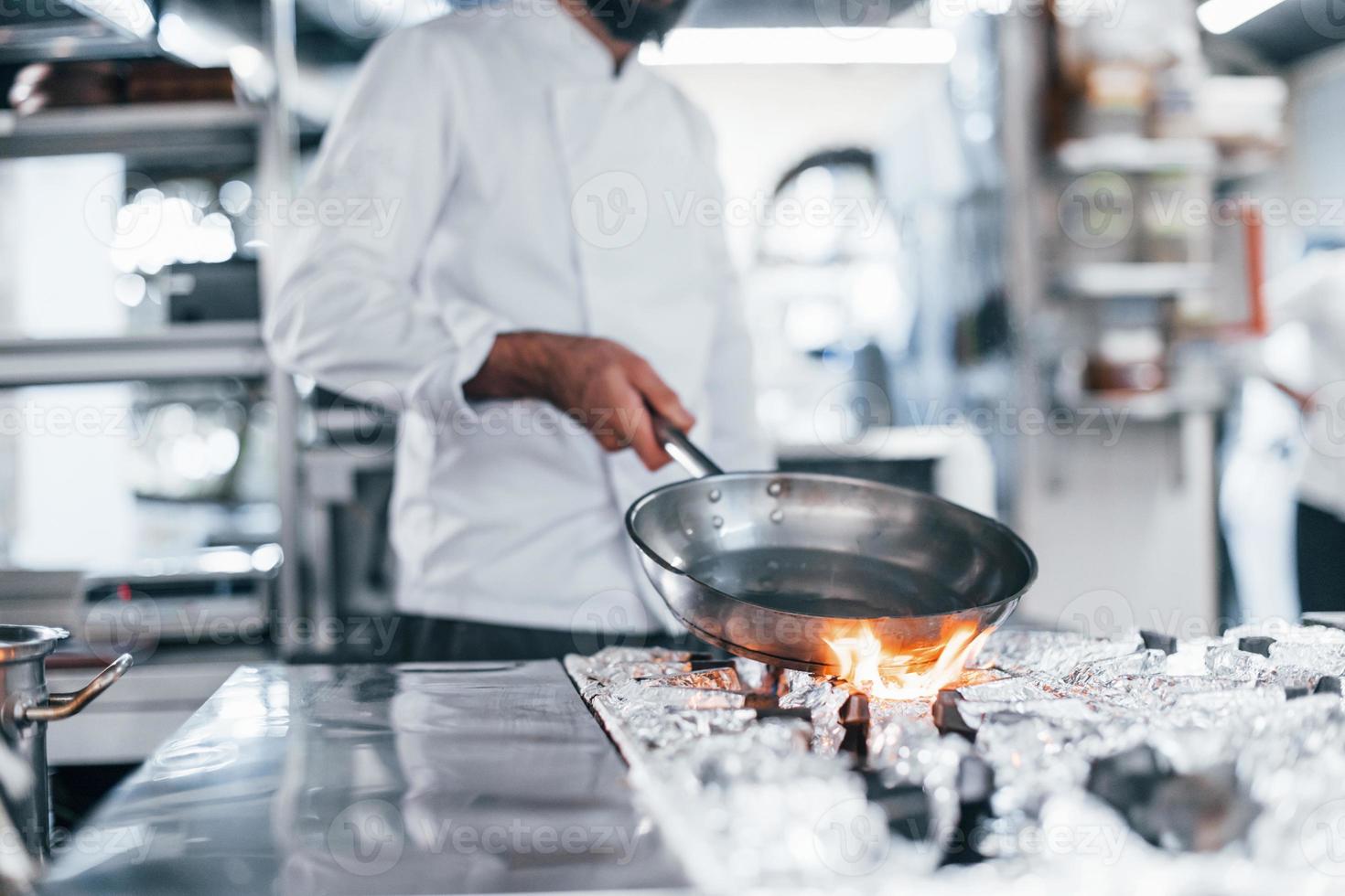 Frying pan is on fire. Chef in white uniform cooking food at kitchen. Busy day at work photo