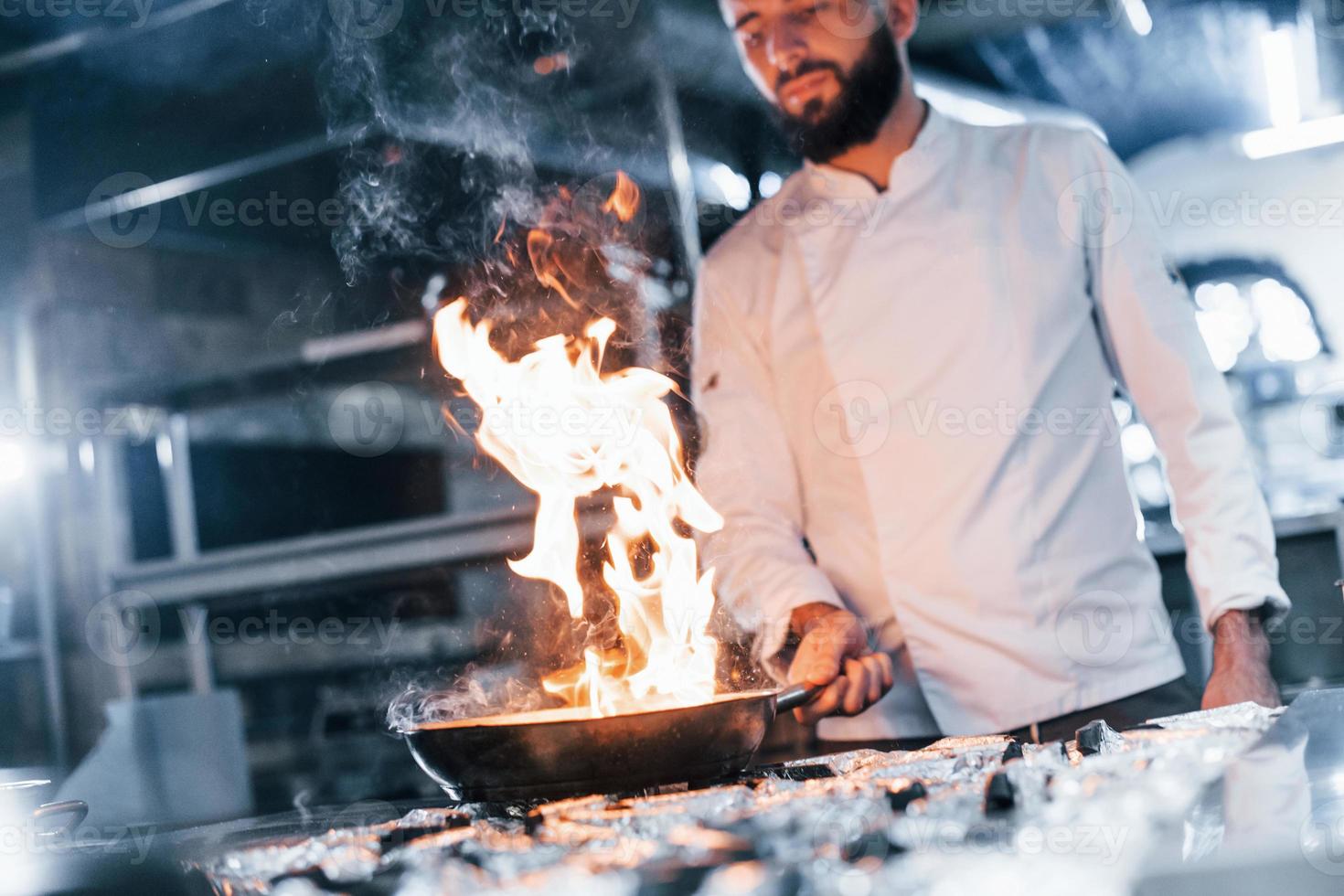 Uses frying pan. Chef in white uniform cooking food at kitchen. Busy day at work photo