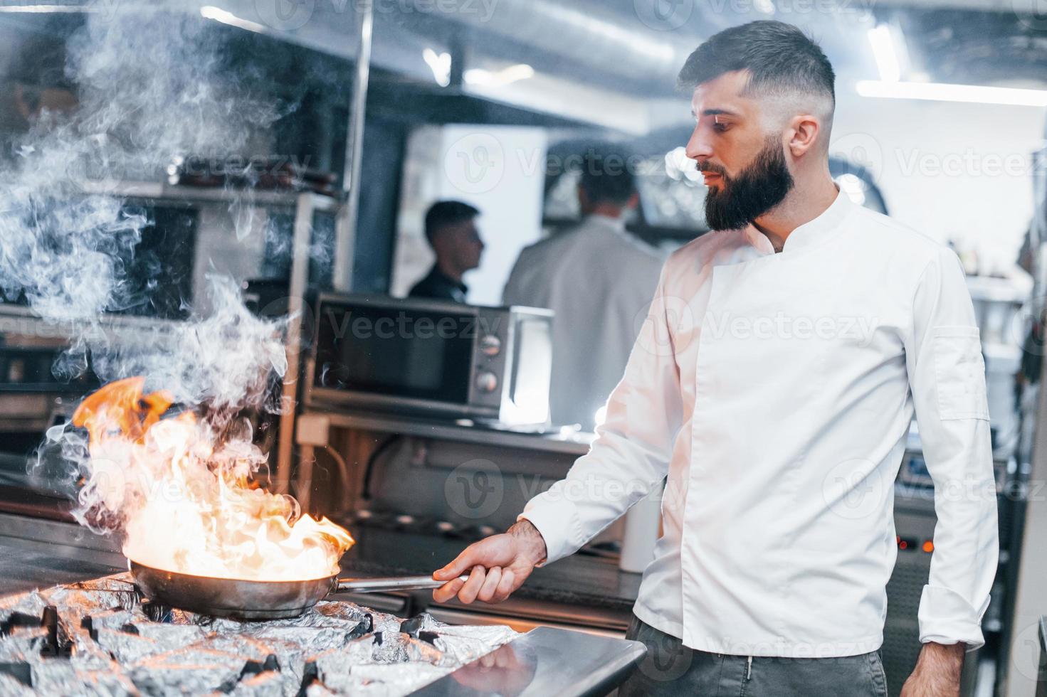 Frying pan is on fire. Chef in white uniform cooking food at kitchen. Busy day at work photo