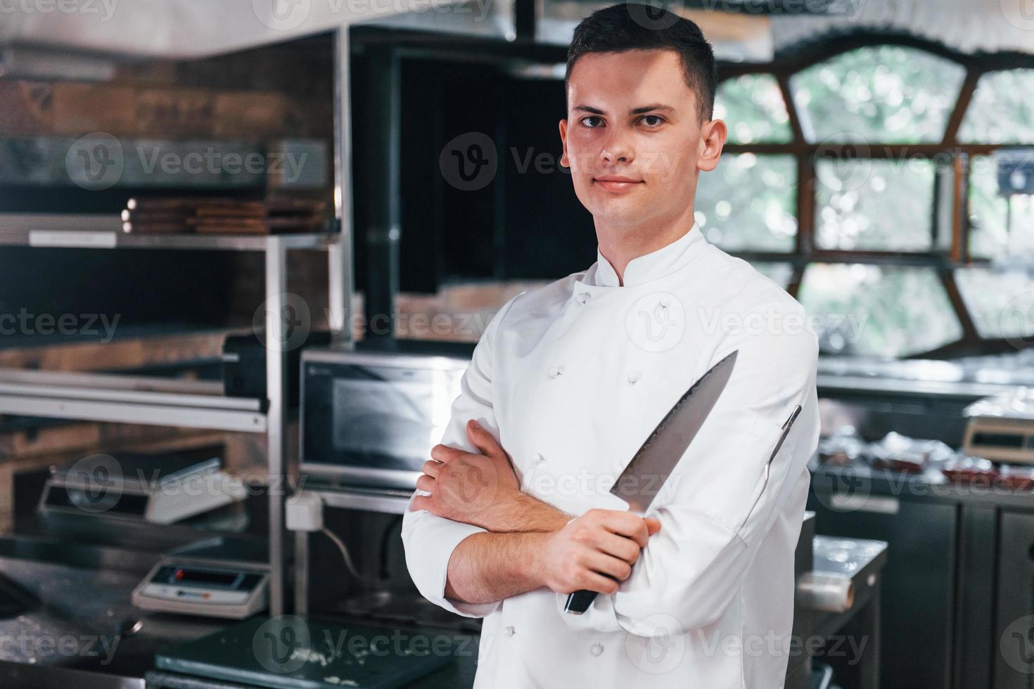 Chef in white uniform standing at kitchen. Holding knife in hands photo