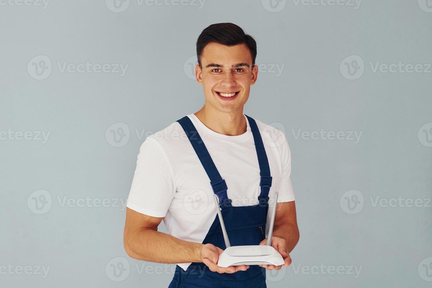 Holds wifi router. Male worker in blue uniform standing inside of studio against white background photo