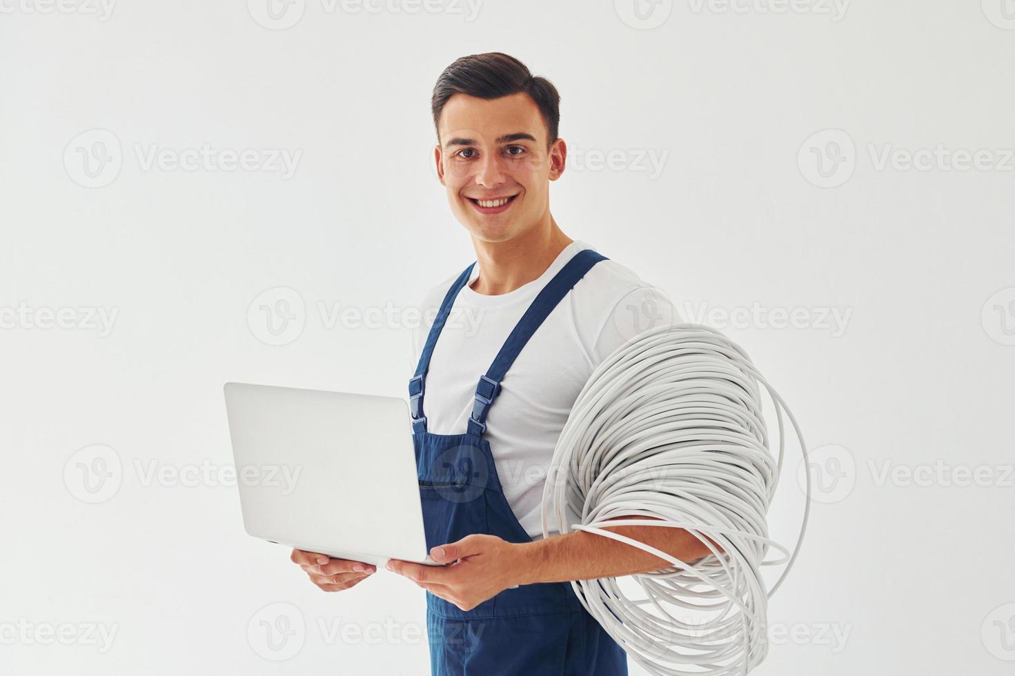 Uses laptop. Male worker in blue uniform standing inside of studio against white background photo