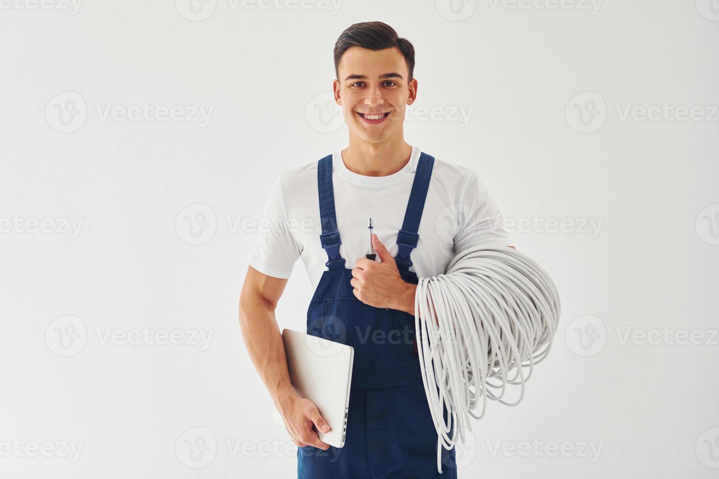 Holds cables, notepad and screwdriver. Male worker in blue uniform standing inside of studio against white background photo