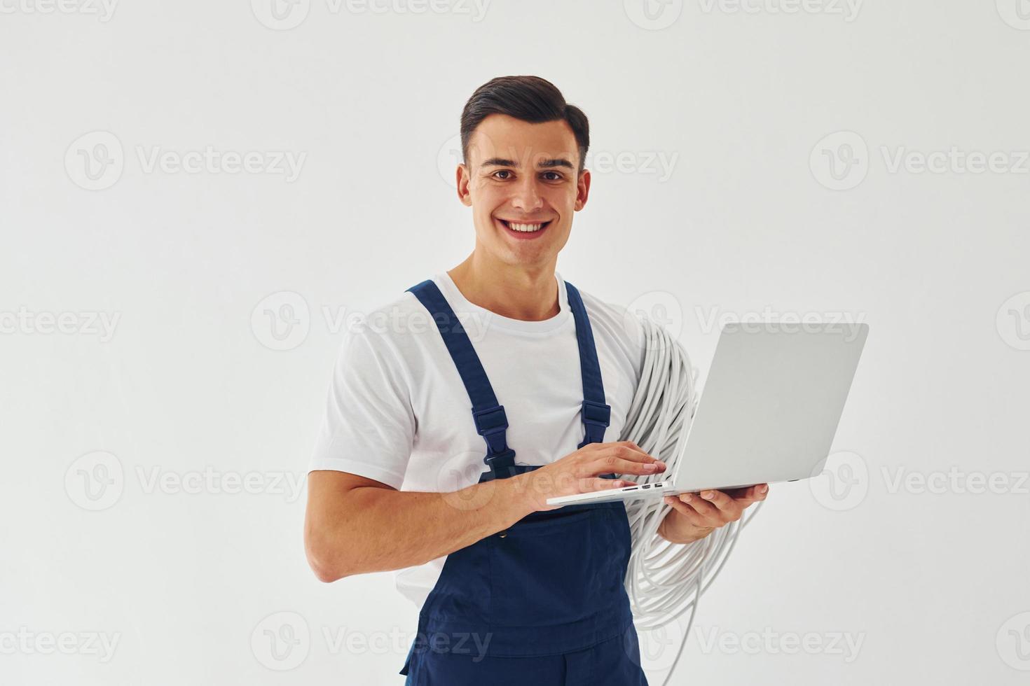 utiliza portátil. trabajador de sexo masculino en uniforme azul de pie dentro del estudio contra el fondo blanco foto