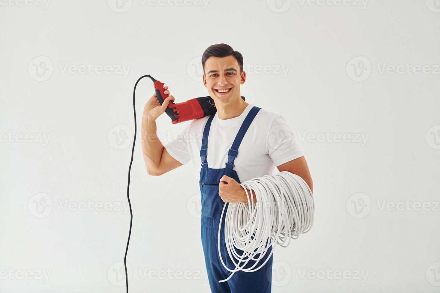 With drill and cables in hands. Male worker in blue uniform standing inside of studio against white background photo