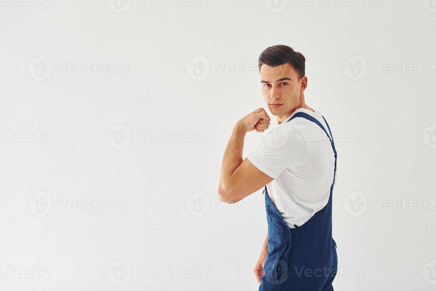 Shows biceps. Ready to work. Male worker in blue uniform standing inside of studio against white background photo
