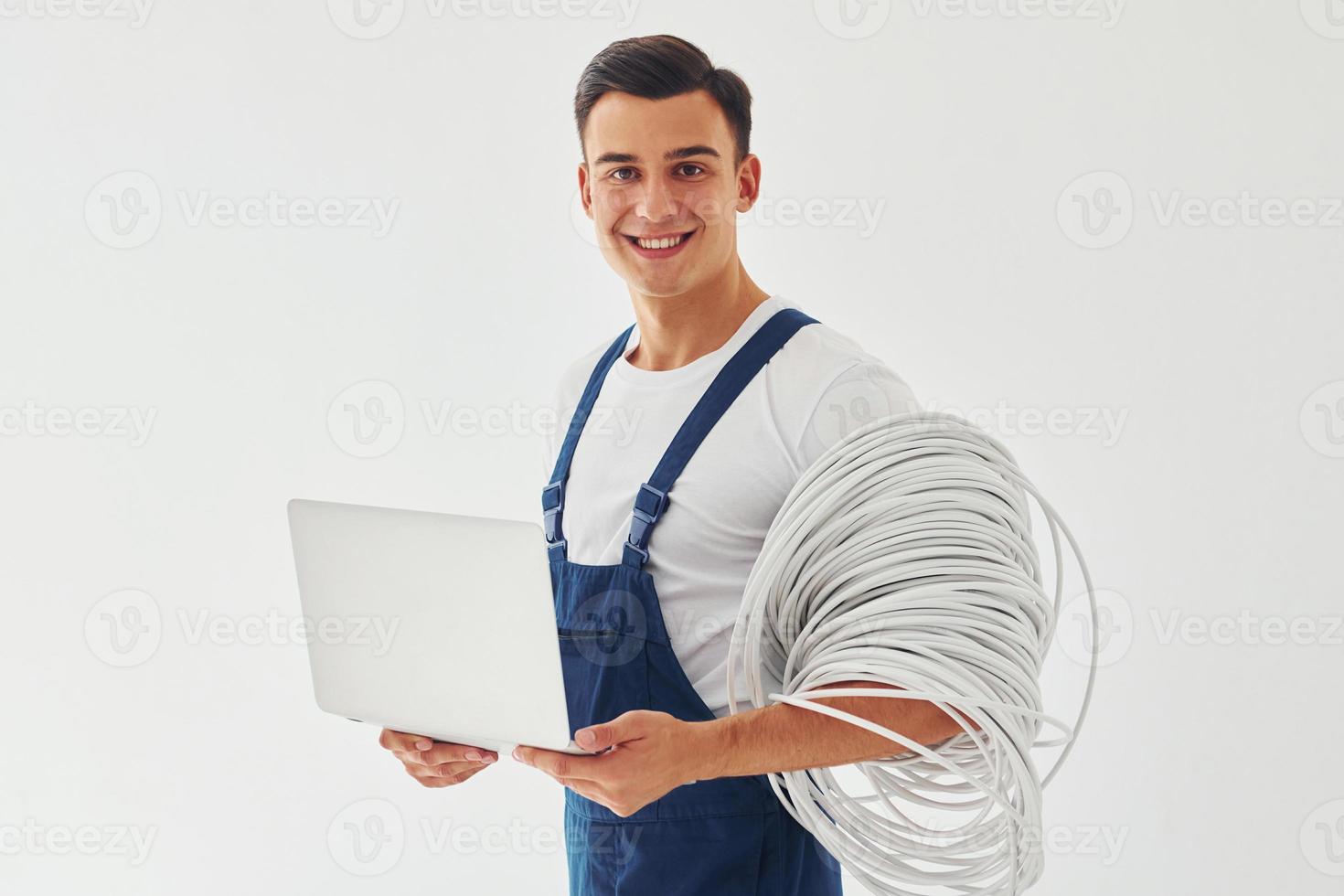 Uses laptop. Male worker in blue uniform standing inside of studio against white background photo
