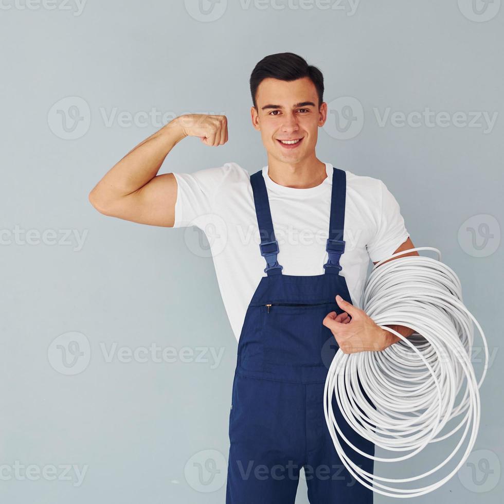 Holds cable. Male worker in blue uniform standing inside of studio against white background photo