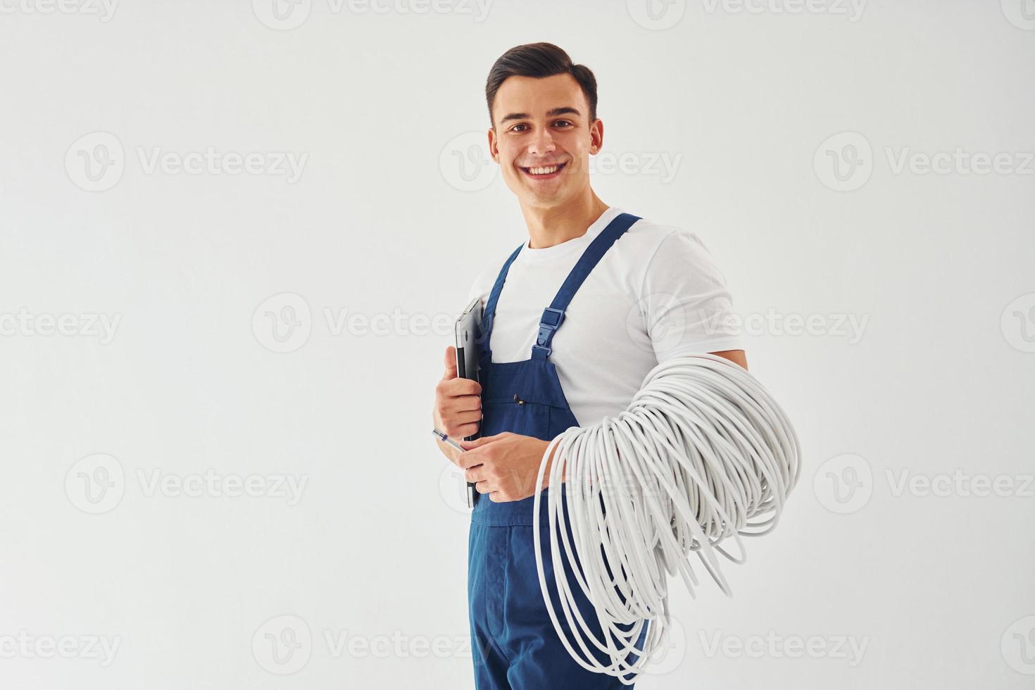 Holds cables, notepad and screwdriver. Male worker in blue uniform standing inside of studio against white background photo