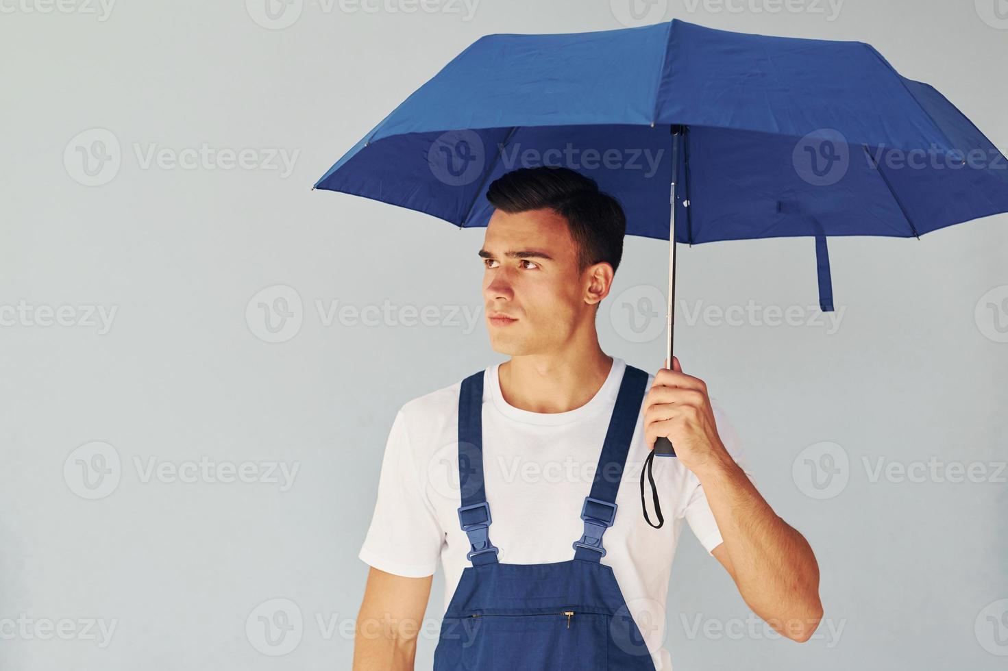 Holds umbrella by hand. Male worker in blue uniform standing inside of studio against white background photo
