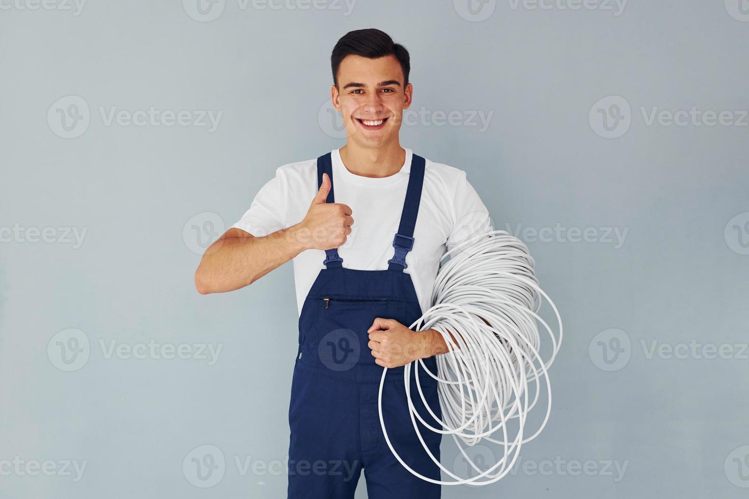 Holds cable. Male worker in blue uniform standing inside of studio against white background photo