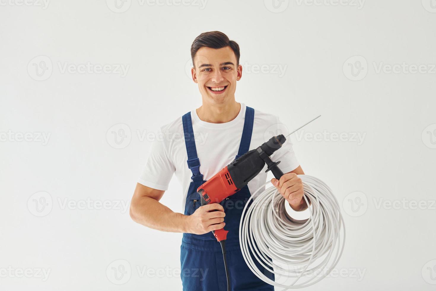 With drill and cables in hands. Male worker in blue uniform standing inside of studio against white background photo