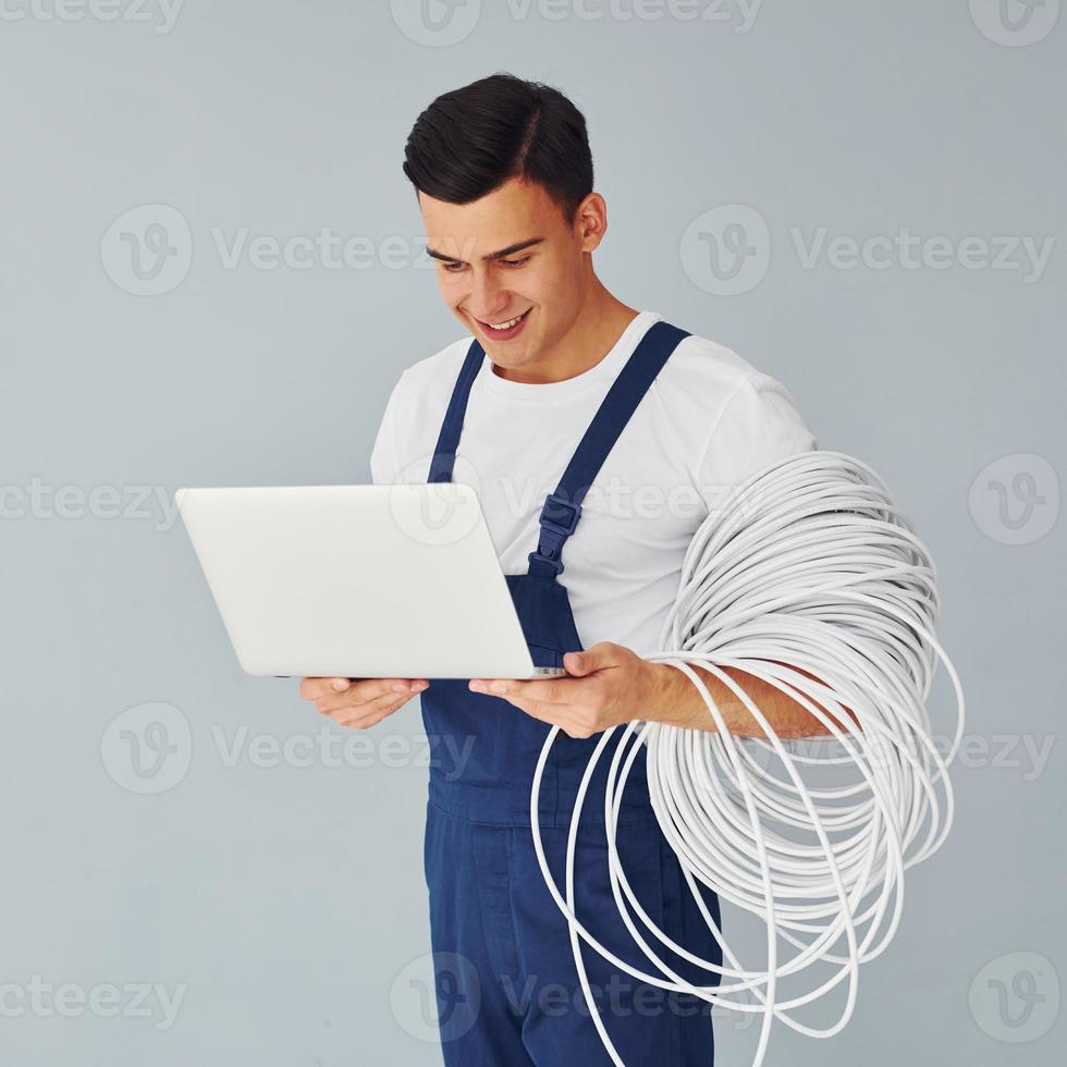 Uses laptop. Male worker in blue uniform standing inside of studio against white background photo